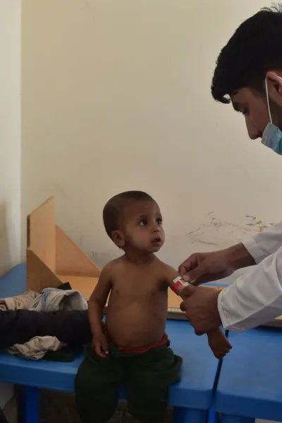 En sjuksköterska undersöker ett barn som sitter på en brits på Läkare Utan Gränsers klinik i Herat. På en stol bredvid sitter en äldre man och ler mot barnet och sjuksköterskan.