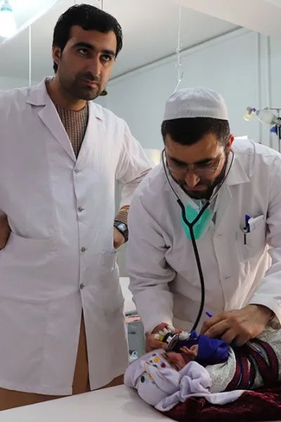 Två män iklädda vita sjukhusrockar står vid en brits på Boost sjukhus, Lashkar Gah, Afghanistan. På britsen ligger en liten bebis.