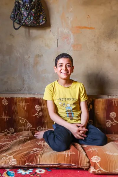 En leende pojke i 10-årsåldern med gul t-shirt sitter på en madrass på golvet i ett kalt rum