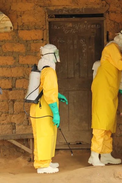 Tre personer i gula dräkter, handskar och masker står vid en dörr till ett teglat hus.