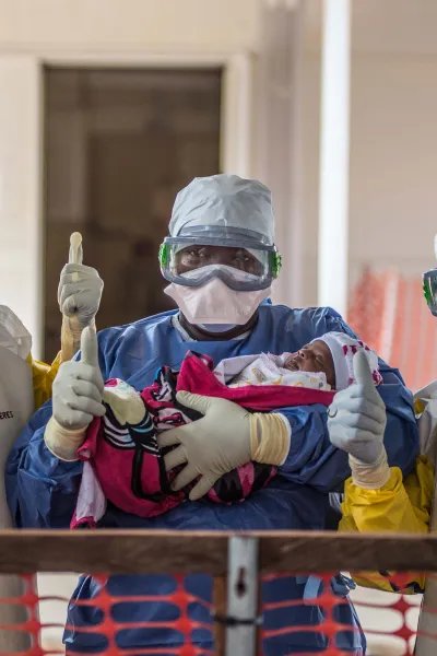 Tre vårdpersonal i skyddsdräkter från vårt ett av våra ebolacentrum gör tummen upp med ett spädbarn i famnen