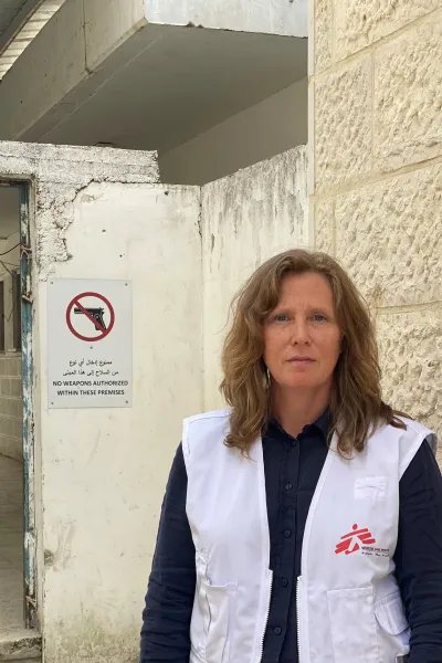 Kvinna tittar in i kameran, iklädd i Läkare Utan Gränser väst, i bakgrunden syns en skylt med vapen och ett sträck över.