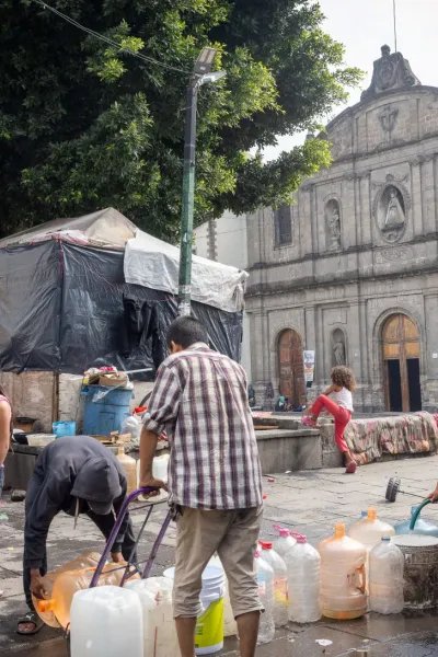 Människor hämtar vatten i stora dunkar på ett torg.