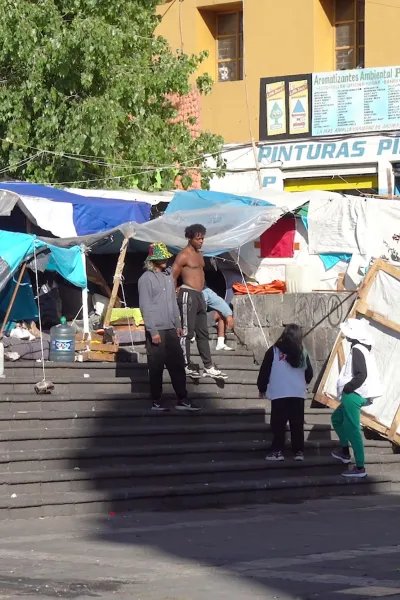 Tältläger i urban miljö. Två personer i Läkare Utan Gränser-västar pratar med en grupp migranter