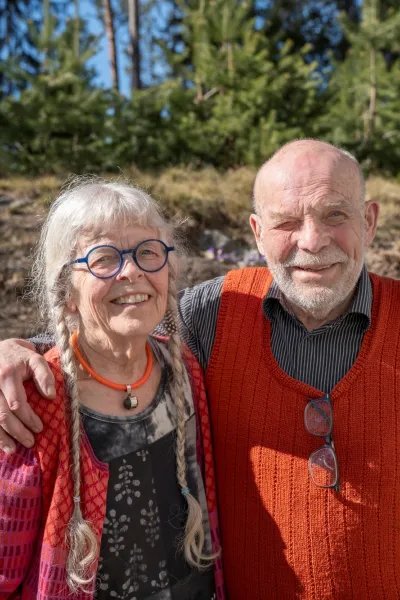 En man och en kvinna står utomhus i solen, mannen håller armen om kvinnans axlar och de tittar in i kameran.