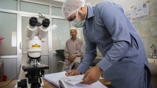 En läkare fyller i en journal. I bakgrunden sitter en patient. 