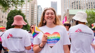 Kvinna med prideflagga och ett hjärta på sin t-shirt med texten Love without borders flankerad av två personer med ryggen till och Läkare utan gränser-västar