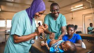 Två personer i kirurggröna kläder undersöker ett barn som ligger på en brits. Bredvid barnet en kvinna som sitter ner vid britsen.