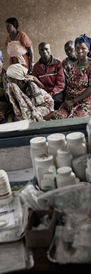 En läkare sitter vid en stor mängd mediciner. Människor väntar utanför ett fönster.