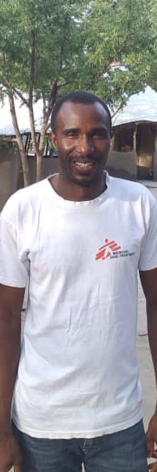 En man står framför några hus i Sydsudan och tittar in i kameran.