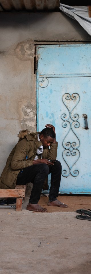 17-åriga Hassan från Sudan sitter på träbrits utanför ett hus i Libyen. 