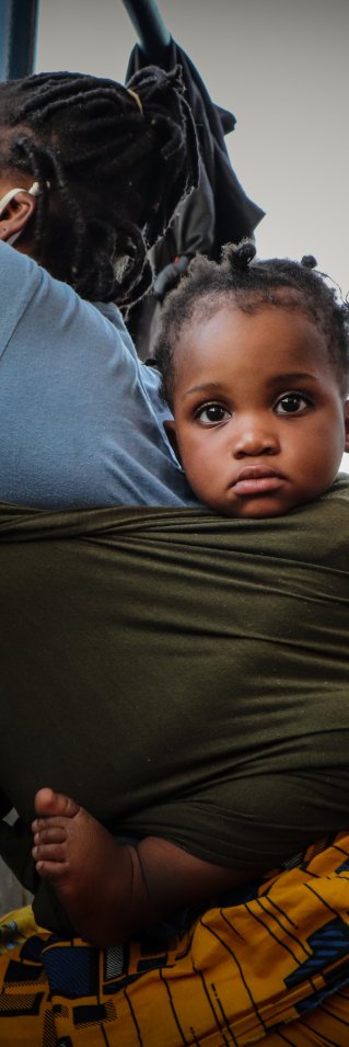 En mamma med ett litet barn i en sjal på ryggen lutar sig mot relingen på en båt och tittar ut över havet