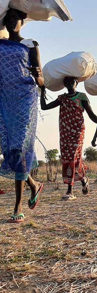 Kvinnor, boende i Riang, Sydsudam, går snett mot kameran på rad. De bär på förnödenheter som ska delas ut till boende i området.