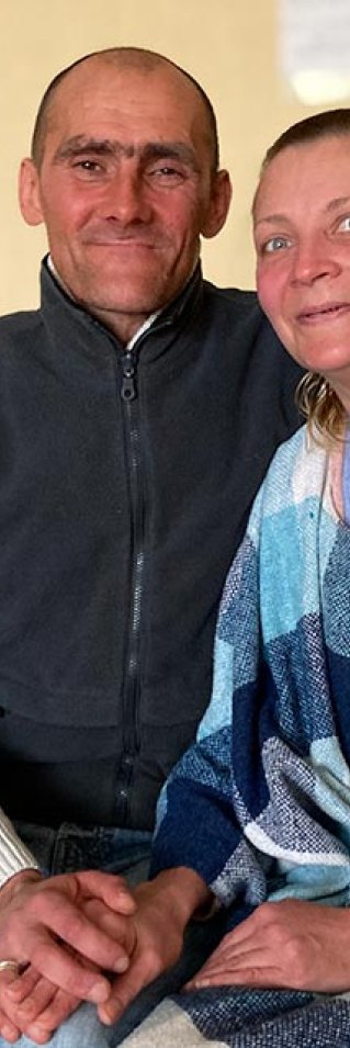 Sergej och Natasha sitter på en säng och håller varandras händer. 