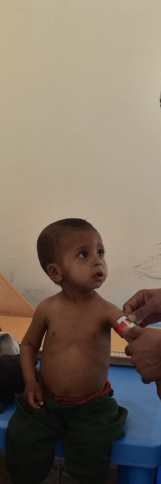 En sjuksköterska undersöker ett barn som sitter på en brits på Läkare Utan Gränsers klinik i Herat. På en stol bredvid sitter en äldre man och ler mot barnet och sjuksköterskan.