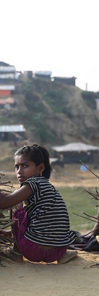 En flicka i flyktinglägret Cox's Bazar, Bangladesh, sitter på huk bredvid en pojke och en hög med grenar och tittar mot kameran.
