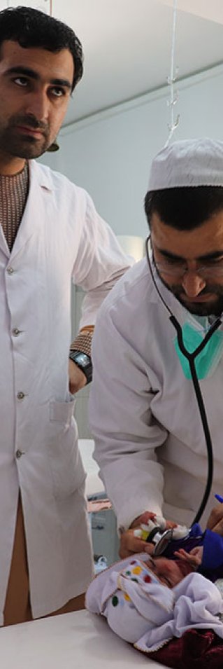 Två män iklädda vita sjukhusrockar står vid en brits på Boost sjukhus, Lashkar Gah, Afghanistan. På britsen ligger en liten bebis.