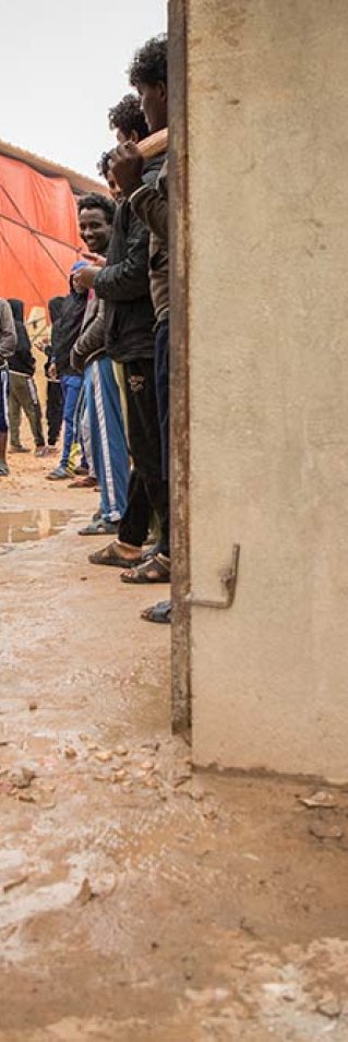 Människor står vid en dörröppning i förvaret Dahr-el-Jebel i Libyen. Det är lerigt på marken.