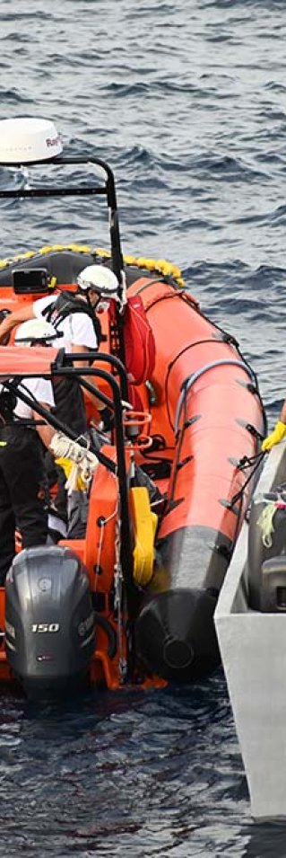 En gummibåt ligger intill en mindre träbåt på centrala Medelhavet. På träbåten ligger flera svarta säckar som två män hjälps åt att lyfta över till gummibåten.