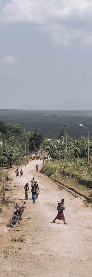 Personer går längs en slutande grusväg. På sidorna syns enklare hus i trä. 