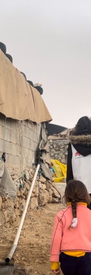 Två personer i Läkare Utan Gränser västar går längs en gatan. Bakom dem går en grupp barn. Husen runt omkring är byggda i betong med tältdukar som tak.