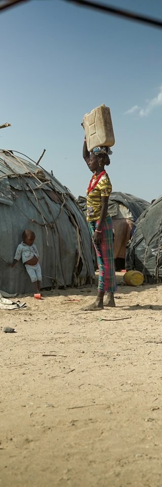 Framför små hyddor står en kvinna hållandes en vattendunk på huvudet.