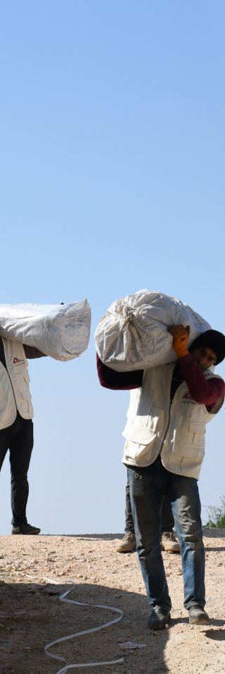 Flera män i Läkare Utan Gränser västar bär påsar med tält över sina axlar.