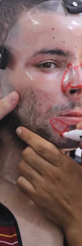 En man har en plastmask för ansiktet. En annan personer målar på masken med en röd penna.