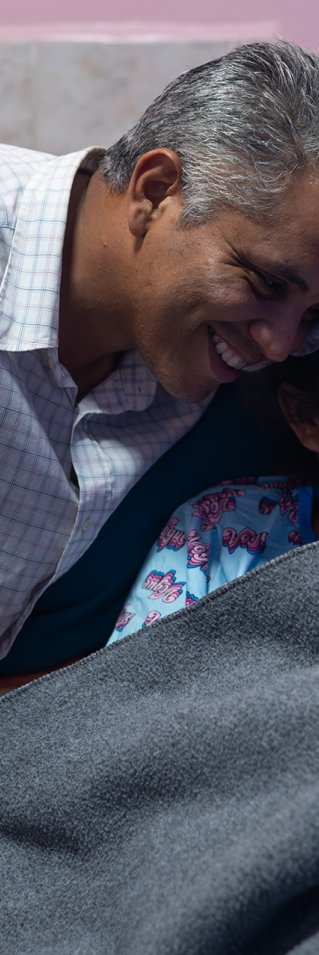 En man håller om en kvinna som ligger i en sjukhussäng. Tillsammans tittar de på ett nyfött barn.