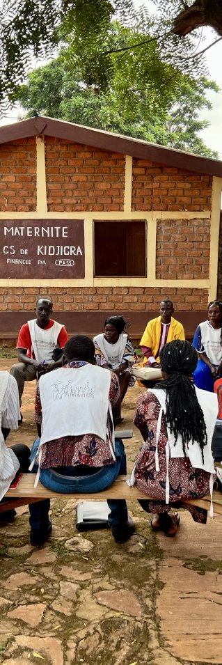 en grupp vårdpersonal på en hälsocentral sitter på träbänkar under ett träd utanför byggnaden