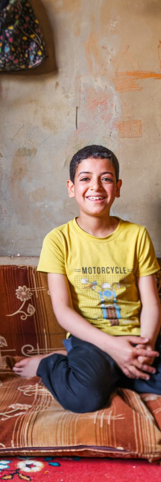 En leende pojke i 10-årsåldern med gul t-shirt sitter på en madrass på golvet i ett kalt rum