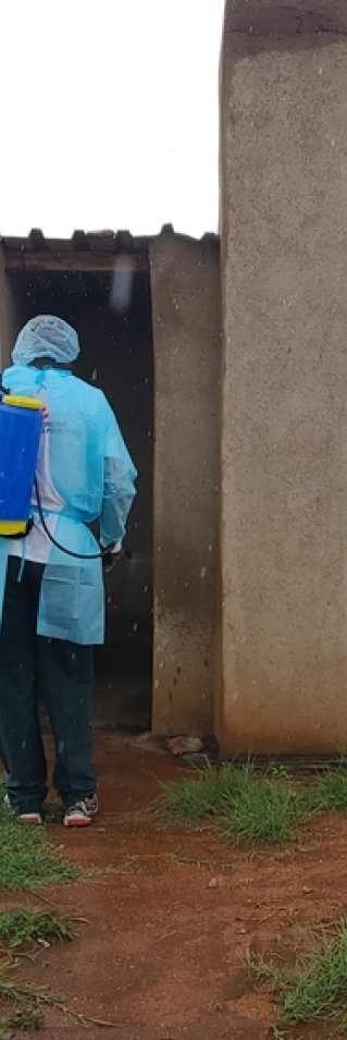 En person med skyddskläder och en container på ryggen står vid en liten betongbyggnad.