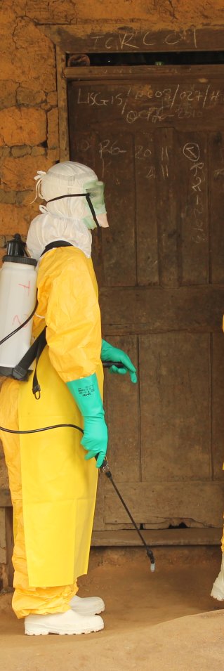 Tre personer i gula dräkter, handskar och masker står vid en dörr till ett teglat hus.