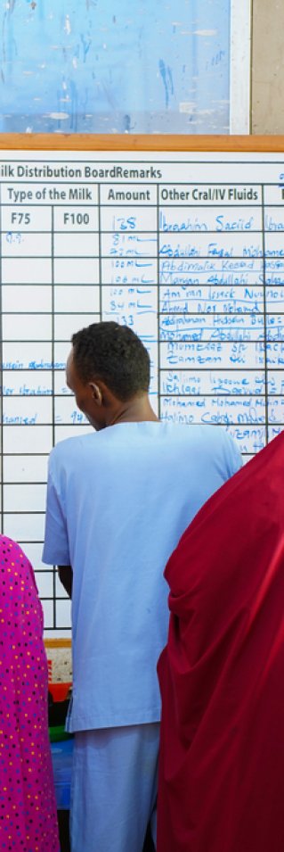 Kvinnor i färglada tyger och en man i blått vända mot en whiteboard med information på.