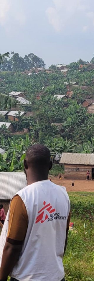 En man i Läkare Utan Gränser-väst med ryggen vänd mot kameran vänd mot gröna kullar, bananplantor och hus i bakgrunden.