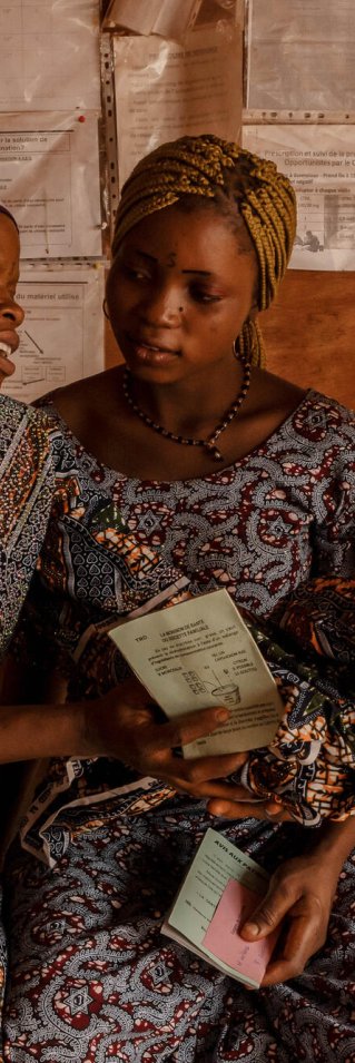 Tre kvinnor i mönstrade klänningar sitter på en bänk. En av kvinnorna räcker över ett spädbarn till en av de andra. 