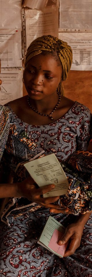 Tre kvinnor i mönstrade klänningar sitter på en bänk. En av kvinnorna räcker över ett spädbarn till en av de andra. 