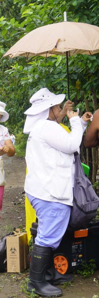 En kvinna i vitt håller upp ett paraply över en man med ryggen mot kameran. Bredvid dem en kvinna i vitt som skriver i en anteckningsbok.