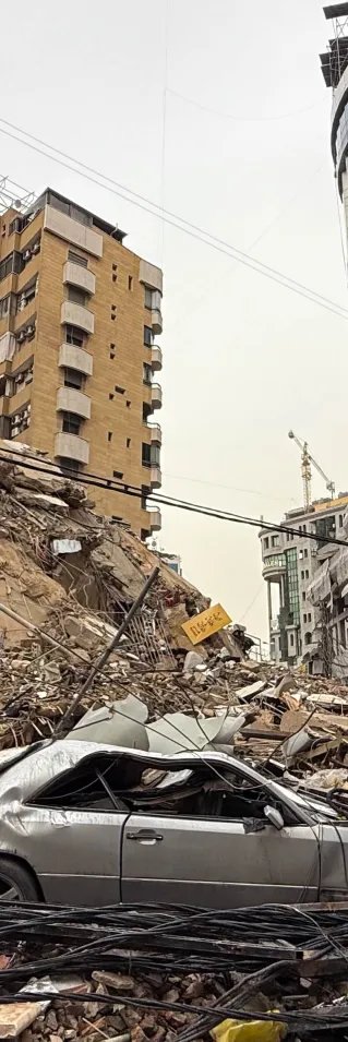 Ett stadsområde med kollapsade byggnader, där en krossad bil och stora rasmassor täcker gatan.