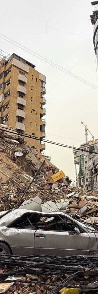 Ett stadsområde med kollapsade byggnader, där en krossad bil och stora rasmassor täcker gatan.