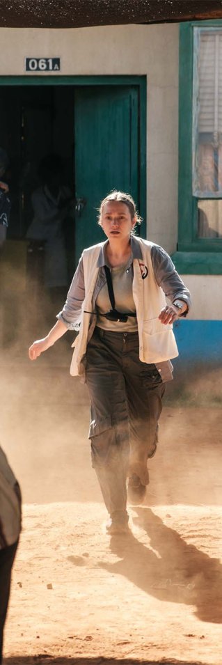 Kvinna i läkarväst srpinger ut ur ett hus och in i en folksamling. Stillbild från filmen Före mörkret