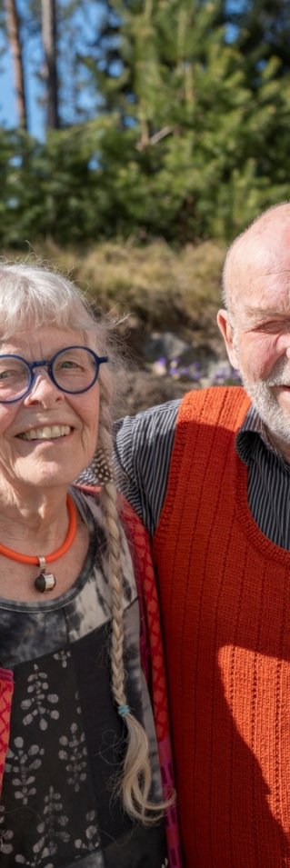 En man och en kvinna står utomhus i solen, mannen håller armen om kvinnans axlar och de tittar in i kameran.