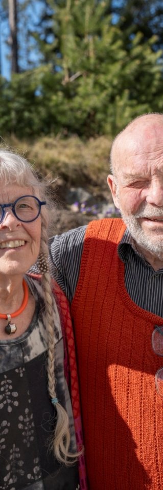 En man och en kvinna står utomhus i solen, mannen håller armen om kvinnans axlar och de tittar in i kameran.