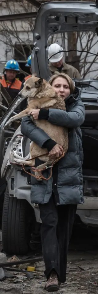 En kvinna håller en hund i famnen bland förstörda hus.