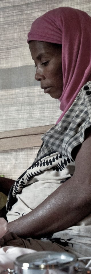 En kvinna med sjal sitter på en enkel säng med ett barn liggandes bredvid henne. Barnet bär en syrgasmask.