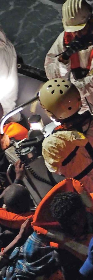En stor grupp människor sitter på en liten gummitbåt i flytvästar mitt i natten medan personal i hjälmar och vita västar försöker hjälpa dem