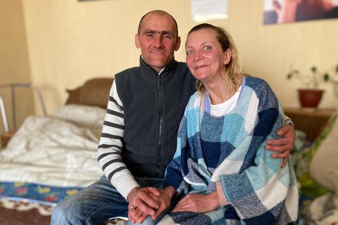 Sergej och Natasha sitter på en säng och håller varandras händer. 