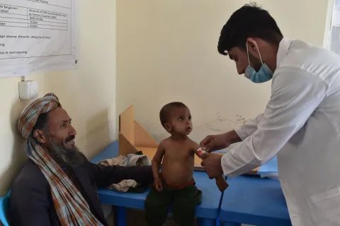En sjuksköterska undersöker ett barn som sitter på en brits på Läkare Utan Gränsers klinik i Herat. På en stol bredvid sitter en äldre man och ler mot barnet och sjuksköterskan.