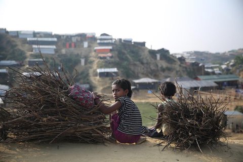 En flicka i flyktinglägret Cox's Bazar, Bangladesh, sitter på huk bredvid en pojke och en hög med grenar och tittar mot kameran.