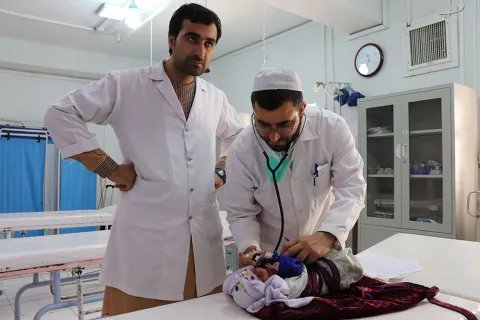 Två män iklädda vita sjukhusrockar står vid en brits på Boost sjukhus, Lashkar Gah, Afghanistan. På britsen ligger en liten bebis.